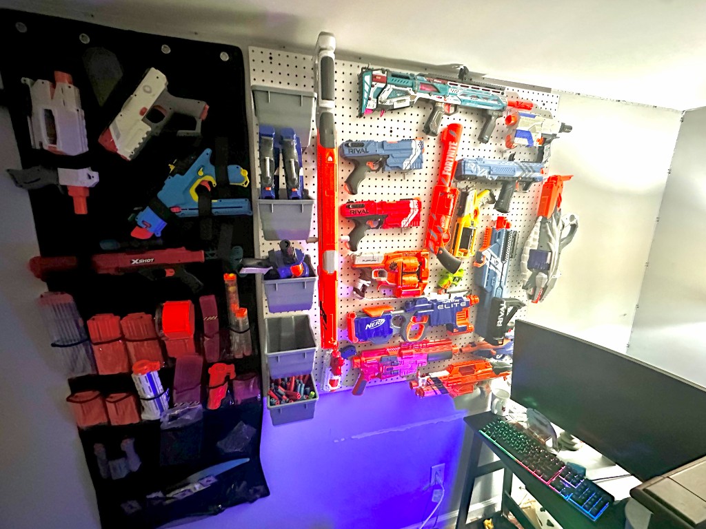 A wall of organized Nerf and foam dart blasters on a white pegboard, with smaller blasters in a black hanging organizer.