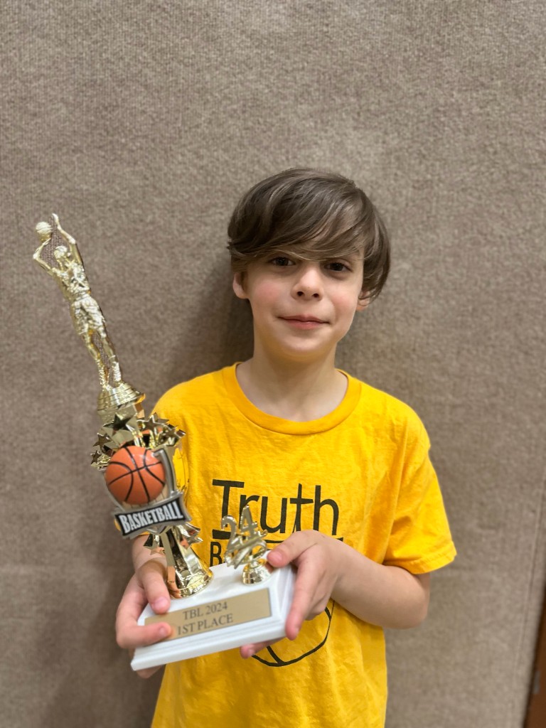 Bastion in a yellow Truth jersey holding a gold basketball trophy with a TBL 2024 1st Place plaque.